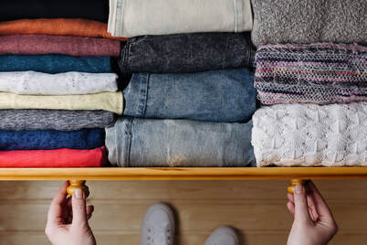Top view of an open drawer organizing colorful folded clothes and jeans next to knit sweaters, with a person's hands holding the drawer handles and their feet visible below.