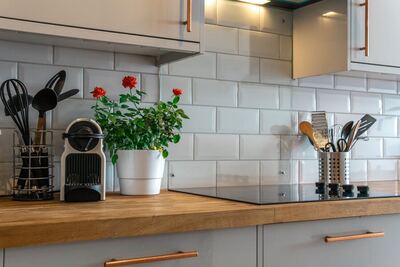 A modern kitchen counter with wooden surface, white subway tile backsplash, and copper handles. On the counter, there's a potted red flowering plant, a holder with black cooking utensils, and a clear container with wooden utensils.